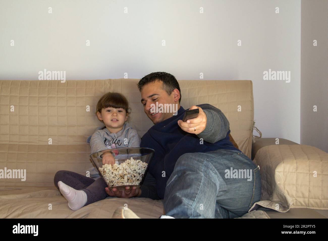 Father and daughter lying on the sofa at home eating popcorn and watching TV. Dad who zaps with the TV remote control. Stock Photo