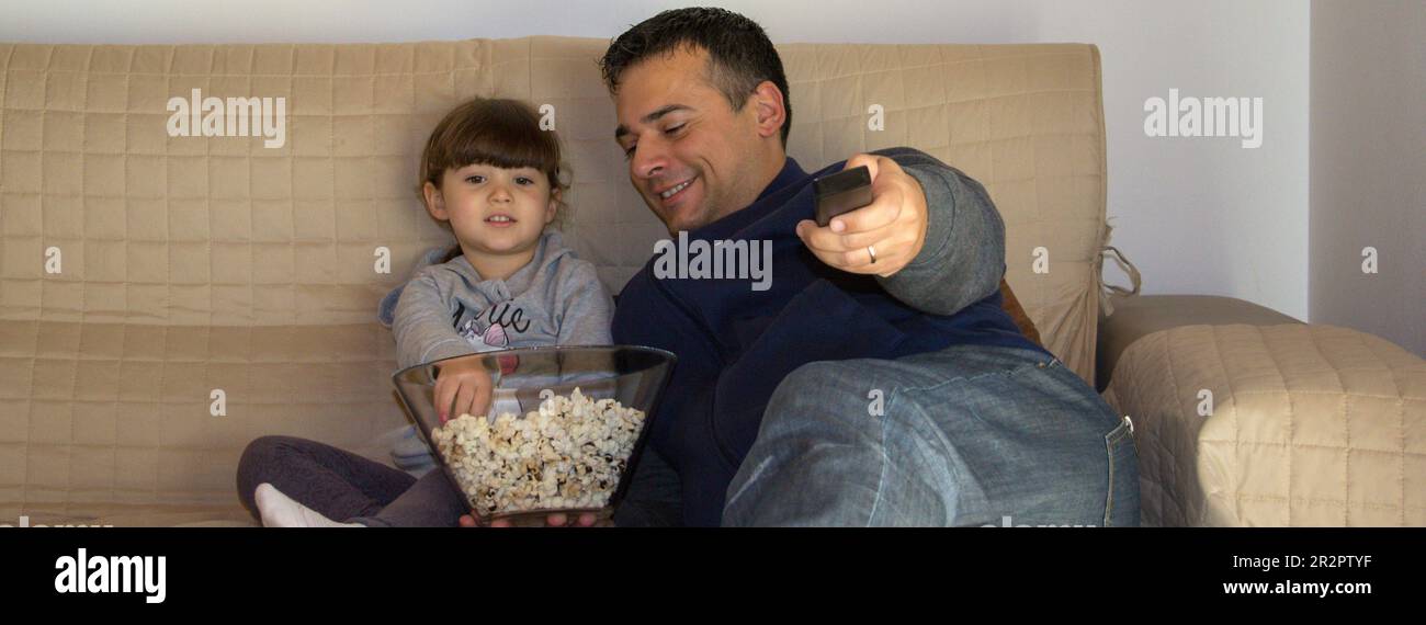 Image of a dad with his daughter lying on the sofa at home while watching TV and eating popcorn. Horizontal banner Stock Photo