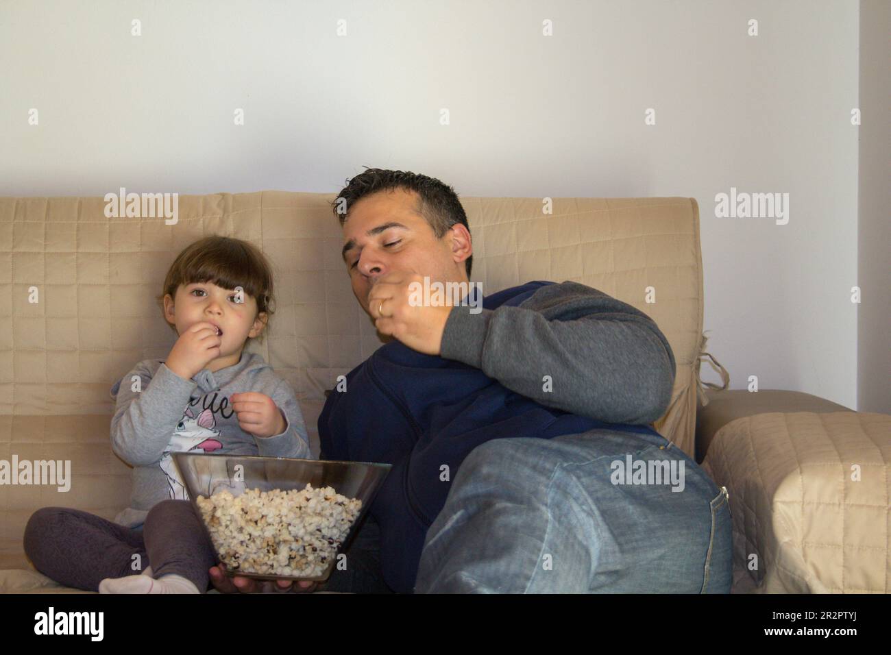 Image of a dad with his daughter lying on the sofa at home while watching TV and eating popcorn. Stock Photo