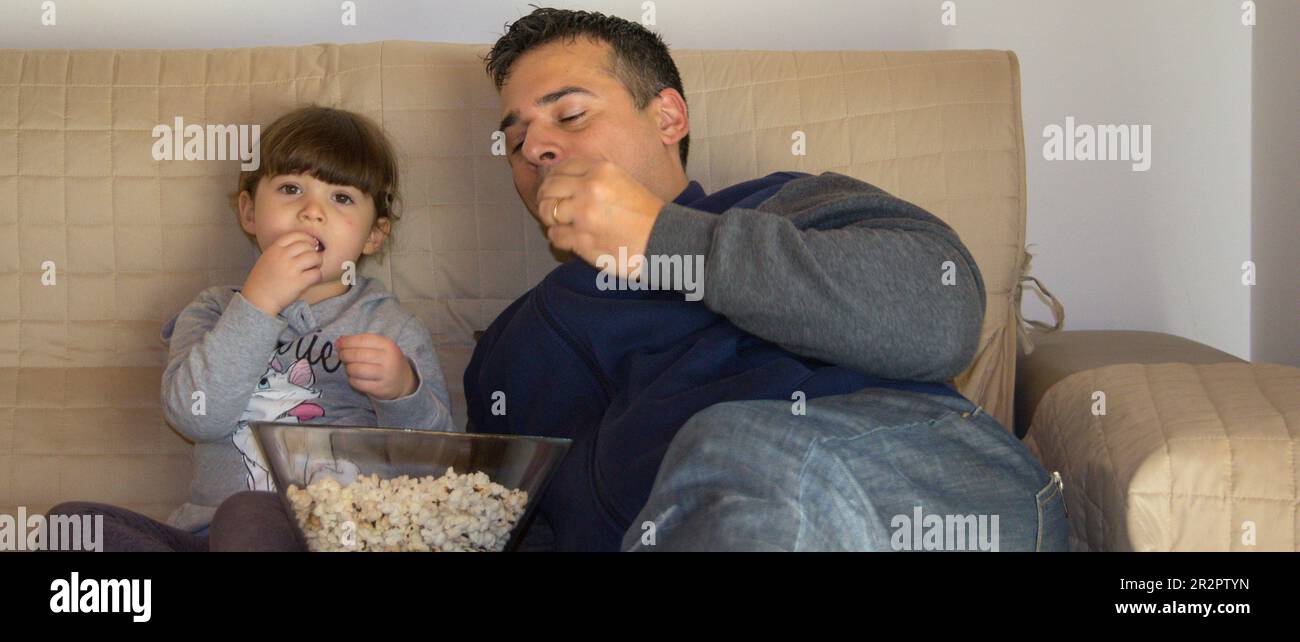 Image of a dad with his daughter lying on the sofa at home while watching TV and eating popcorn. Horizontal banner Stock Photo