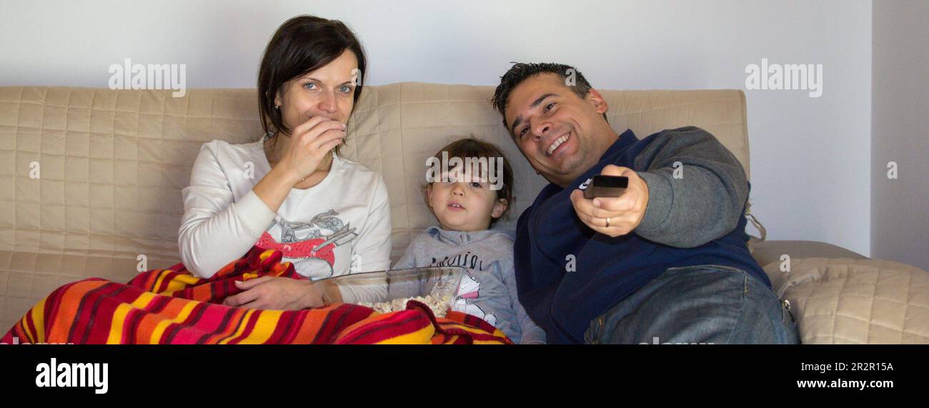 Image of a smiling family lying on the sofa at home watching a movie on TV and eating popcorn. Relaxed family evening. Horizontal banner Stock Photo