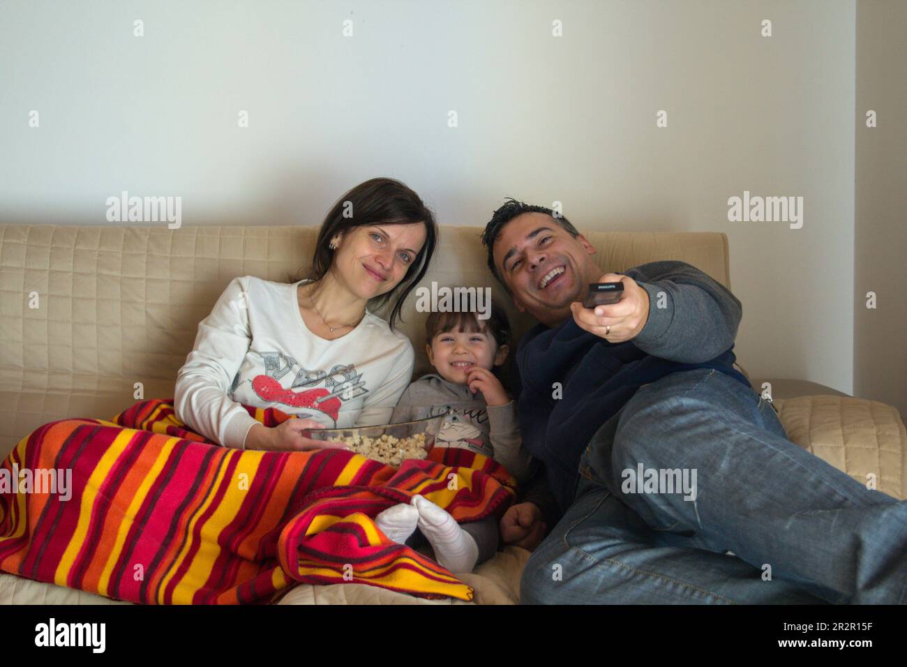 Image of a smiling family lying on the sofa at home watching a movie on TV and eating popcorn. Relaxed family evening Stock Photo