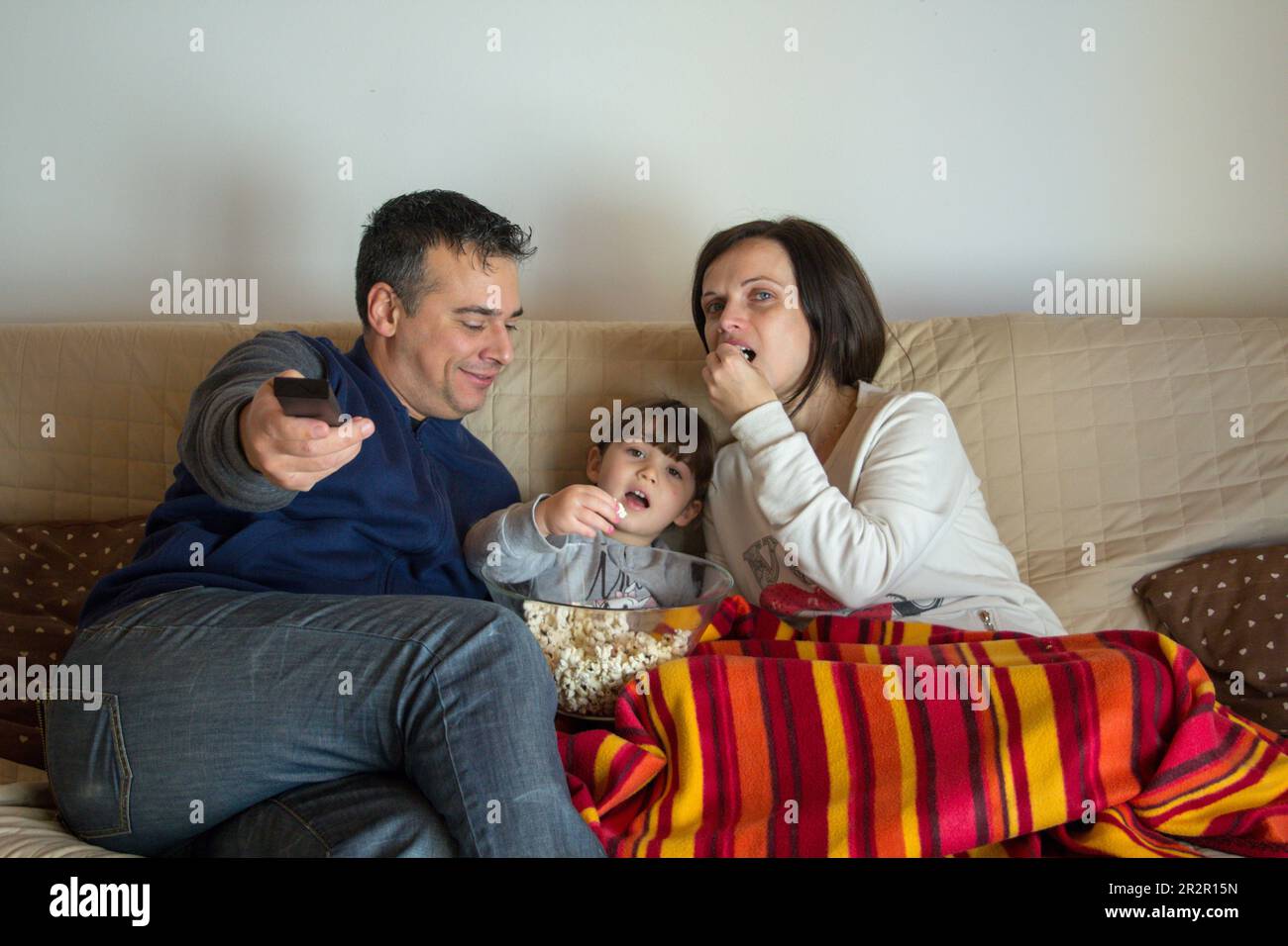 Image of a smiling family lying on the sofa at home watching a movie on TV and eating popcorn. Relaxed family evening Stock Photo