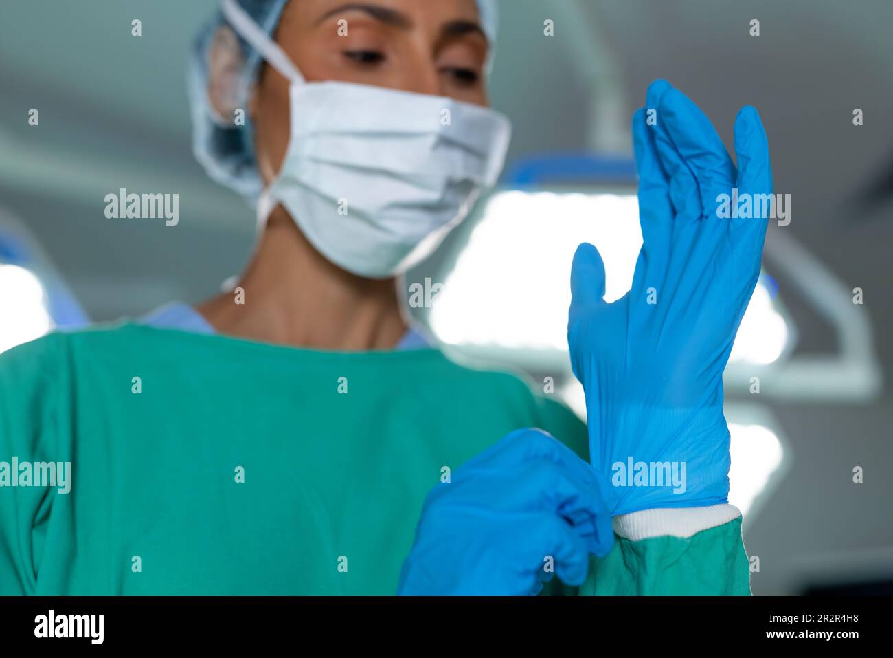 Biracial female surgeon in surgical gown and mask putting on gloves in