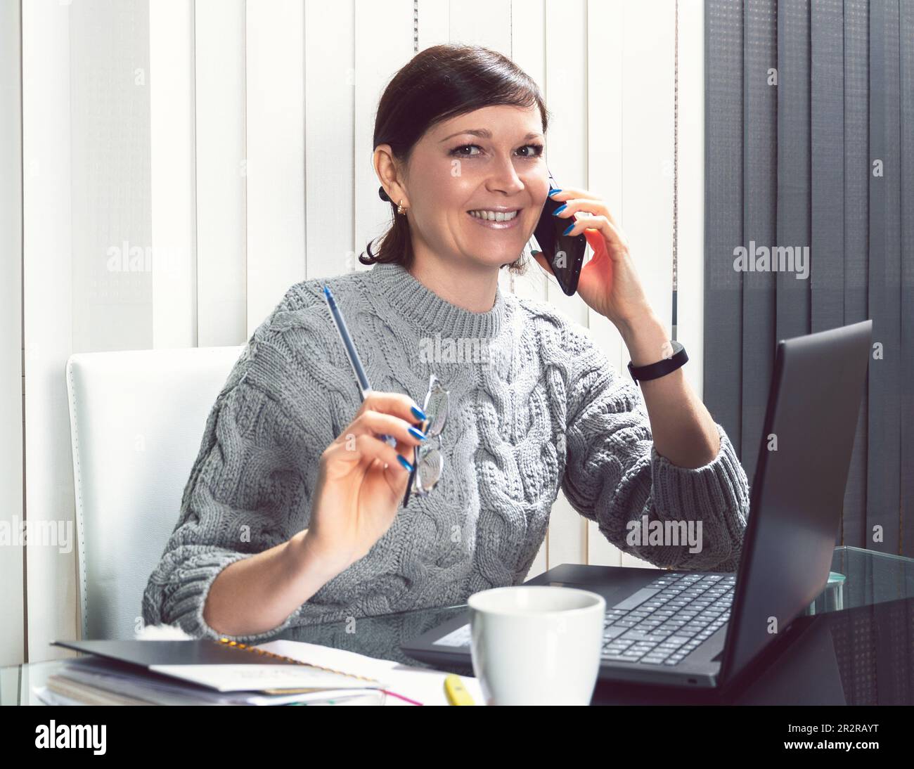 Beautiful young woman with beaming smile, talking on the phone looking at the camera, in front of the laptop, in the modern home office doing business Stock Photo