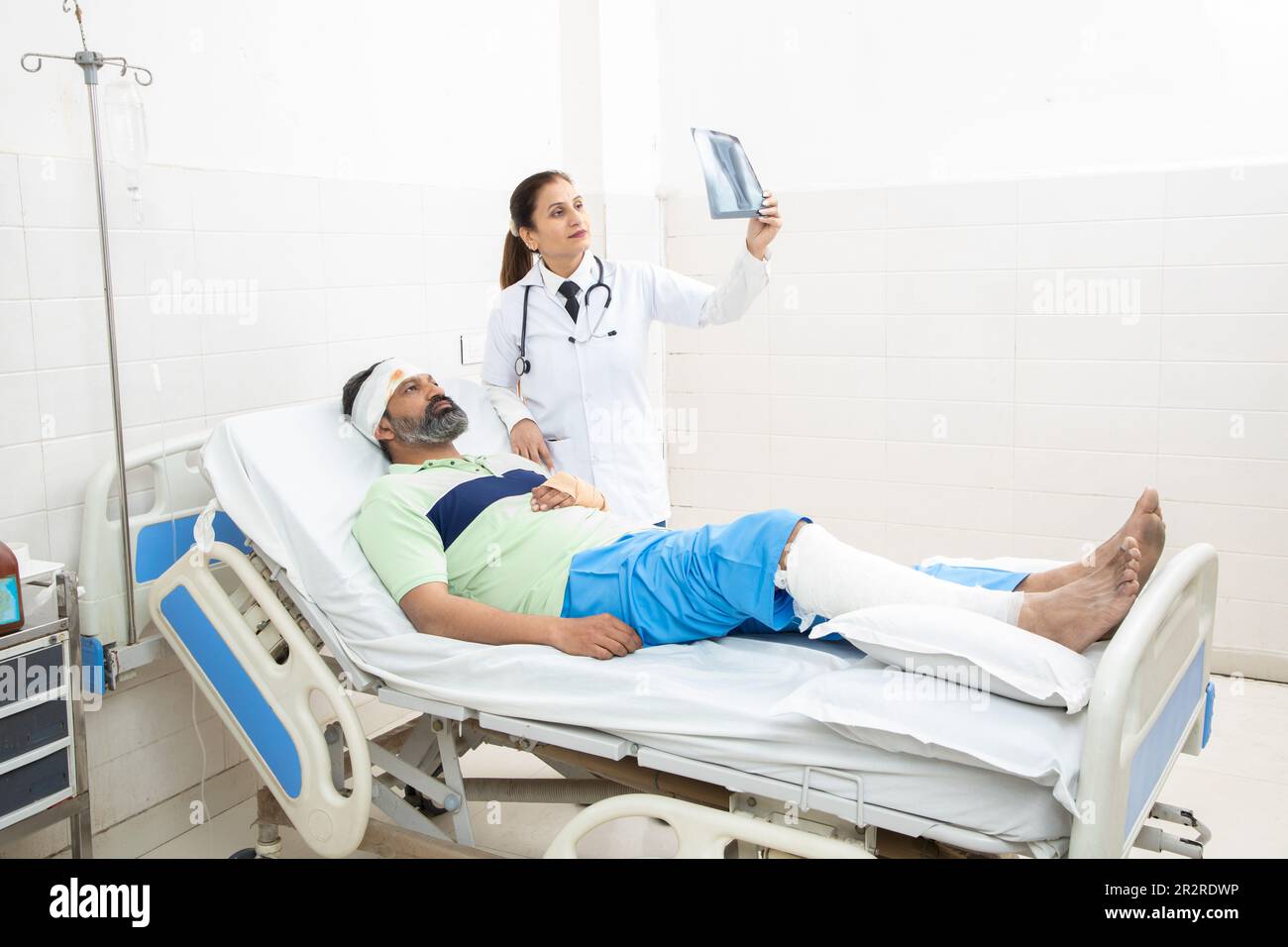 Indian man patient lying on bed with fractured leg and orthopedic doctor looking at foot X-ray film giving recovery advice. Stock Photo