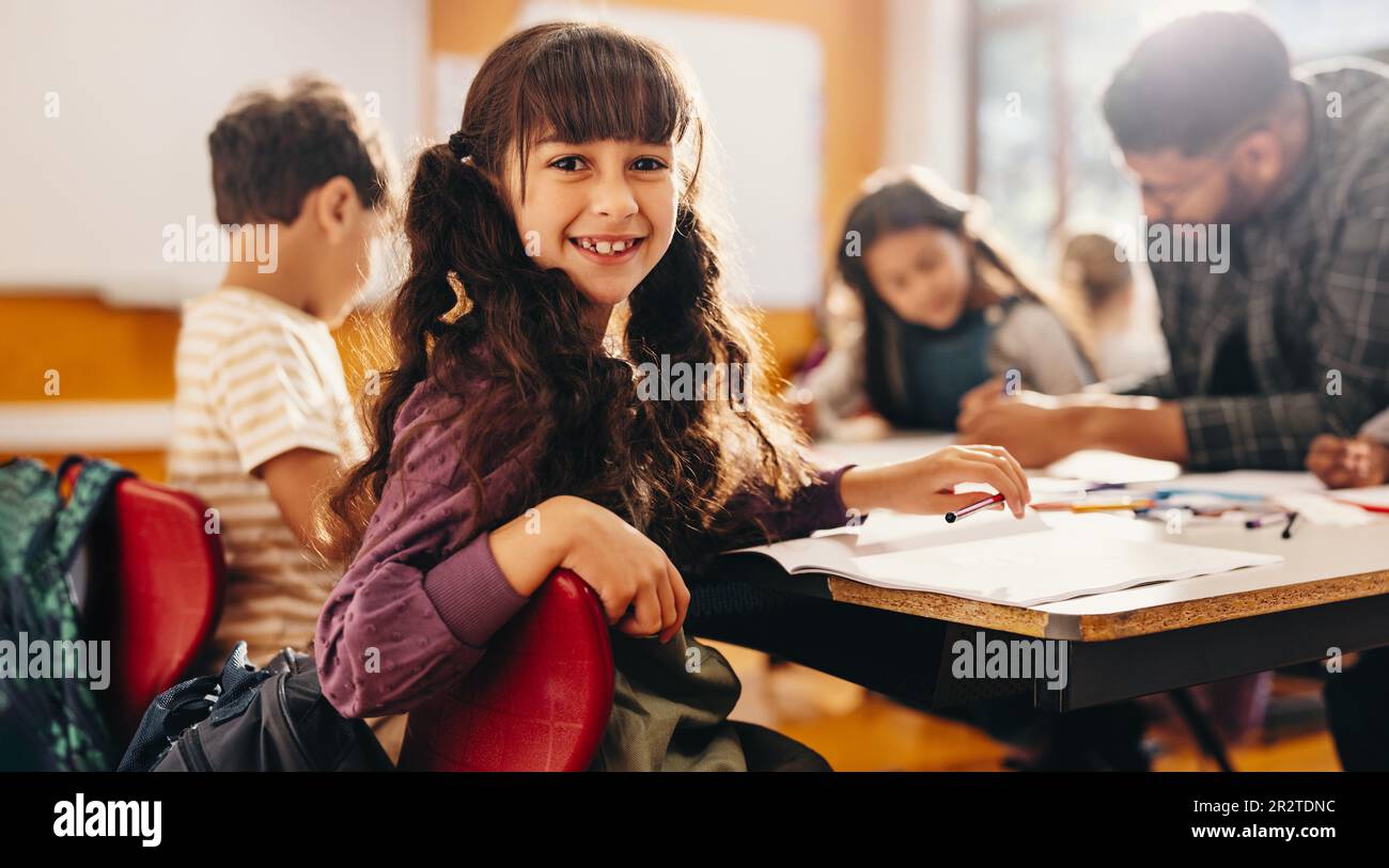 Girl smiling at the camera while sitting in an art and creativity class. Kid enjoying a lesson with her teacher and classmates in elementary school. Stock Photo