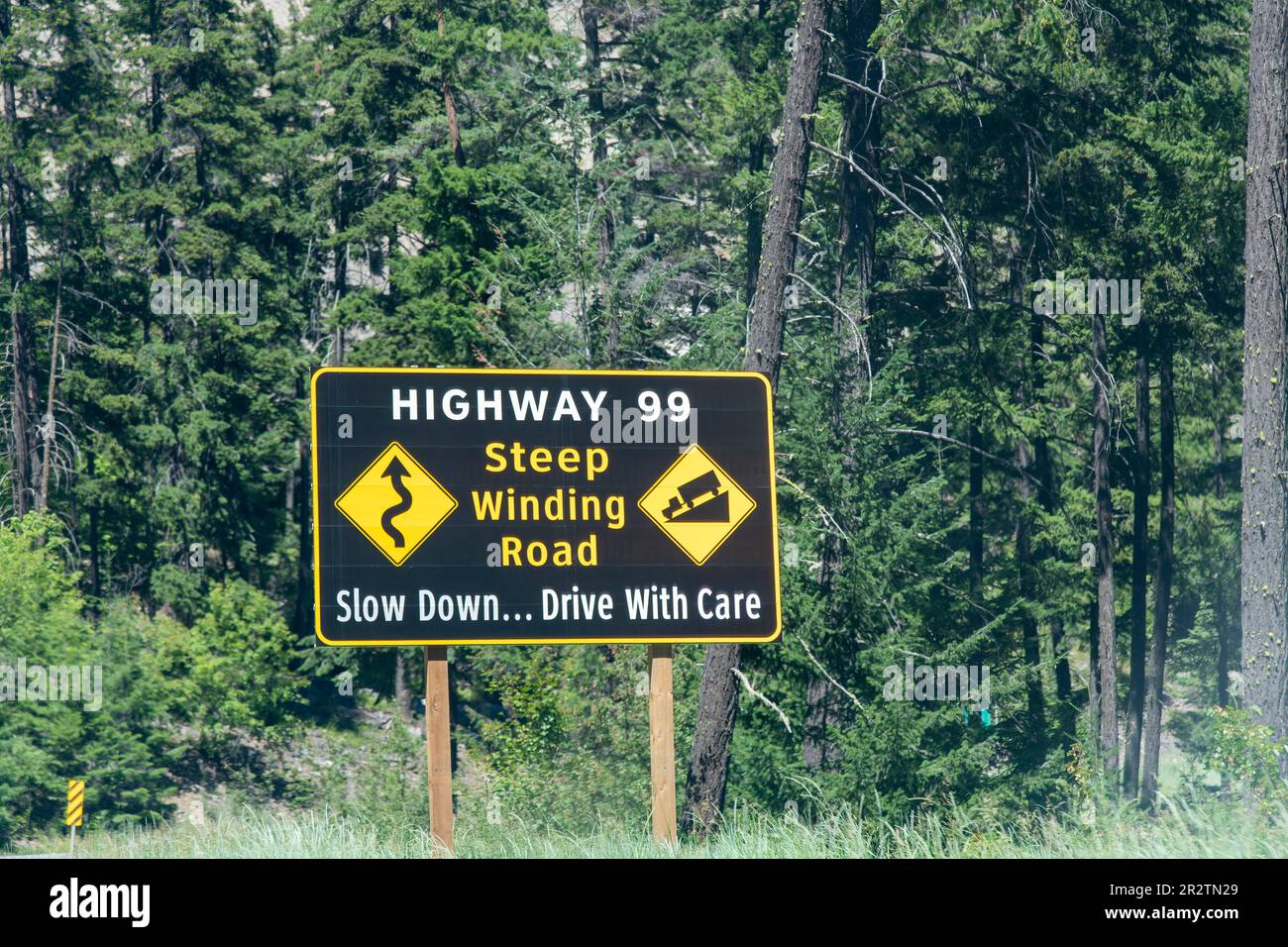 British Columbia, Canada-August 2022; Large yellow road sign between pine trees along Highway 99 warning to slow down because of steep and winding roa Stock Photo