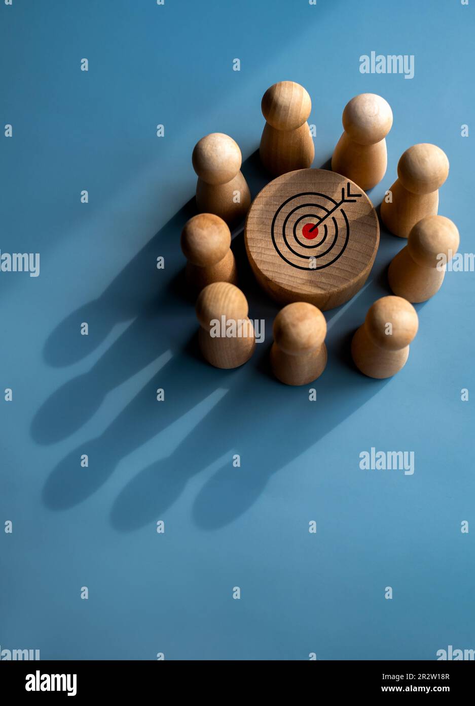 Target icon on round wood block surrounded with small wooden human figures made a crown shape shadow on blue background. Business goal and success, le Stock Photo