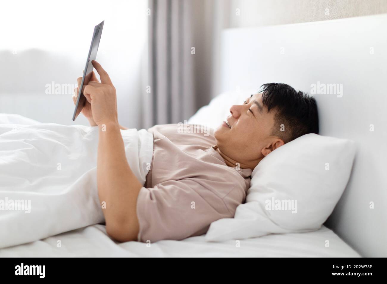 Happy asian man using digital tablet in bed, side view Stock Photo