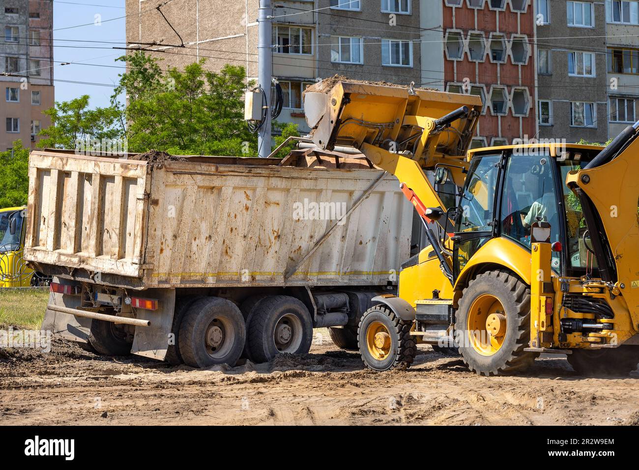 A large tractor bucket pours soil into the back of a large truck at a road construction site. Stock Photo