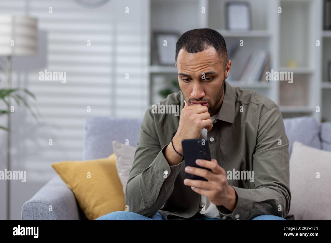 Young african american man sitting on sofa at home and looking at phone upset, received and read bad news, message. Stock Photo
