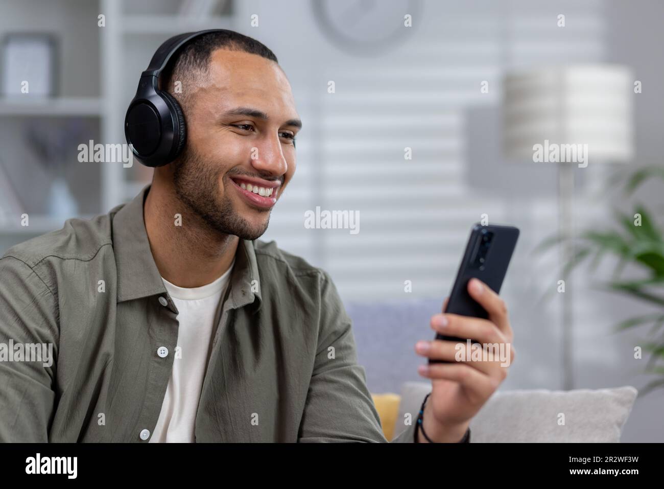 Young Hispanic man sitting at home wearing headphones, holding phone in hand and listening to music, audiobook, podcast, talking on video call. Close- Stock Photo
