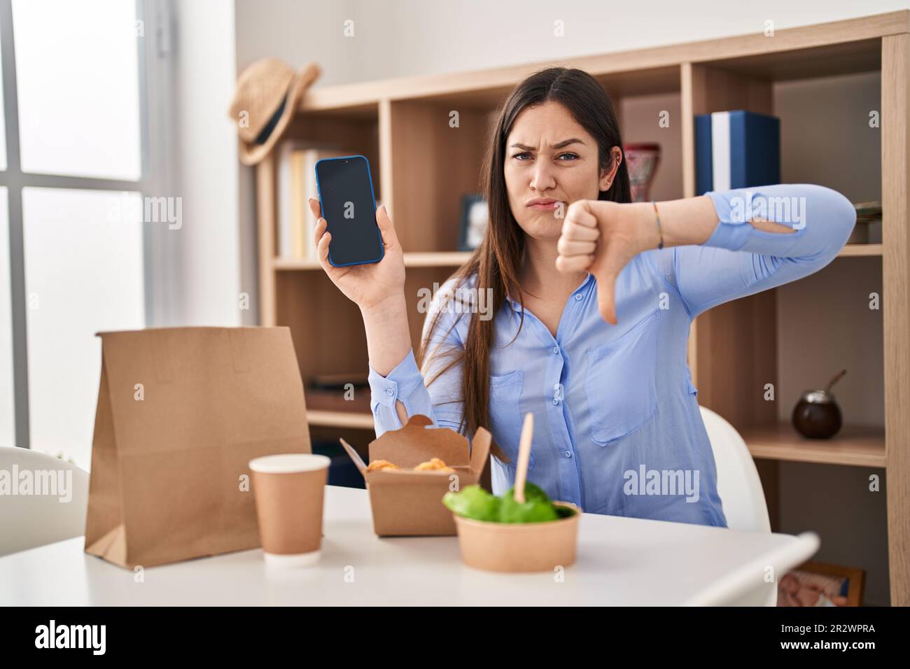 Young brunette woman eating take away food at home showing smartphone ...