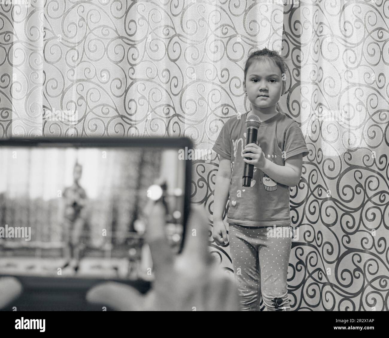 Cute little Asian girl looking at camera holding microphone at home concert. Hands holding tablet for taking video, in unfocus. Lifestyle concept. Bla Stock Photo