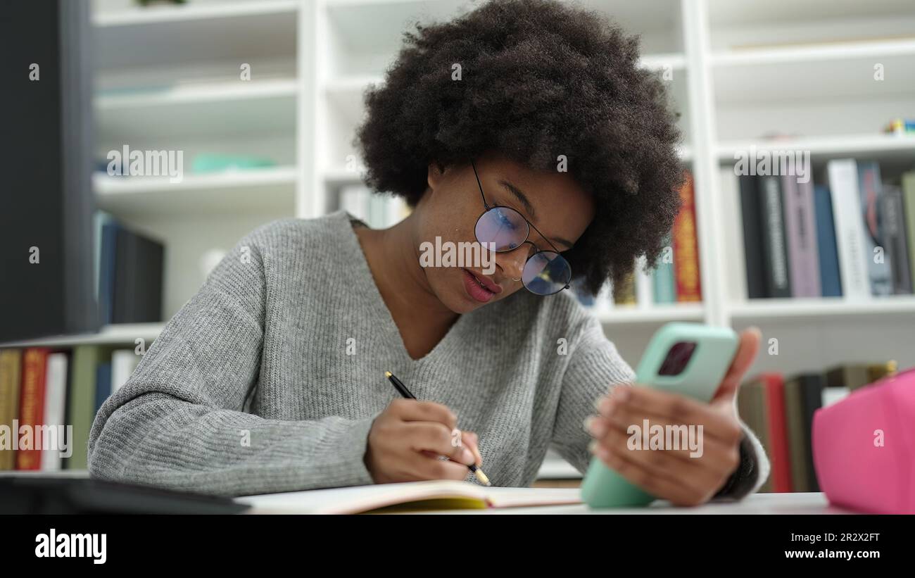 African american woman student using smartphone writing on notebook at ...