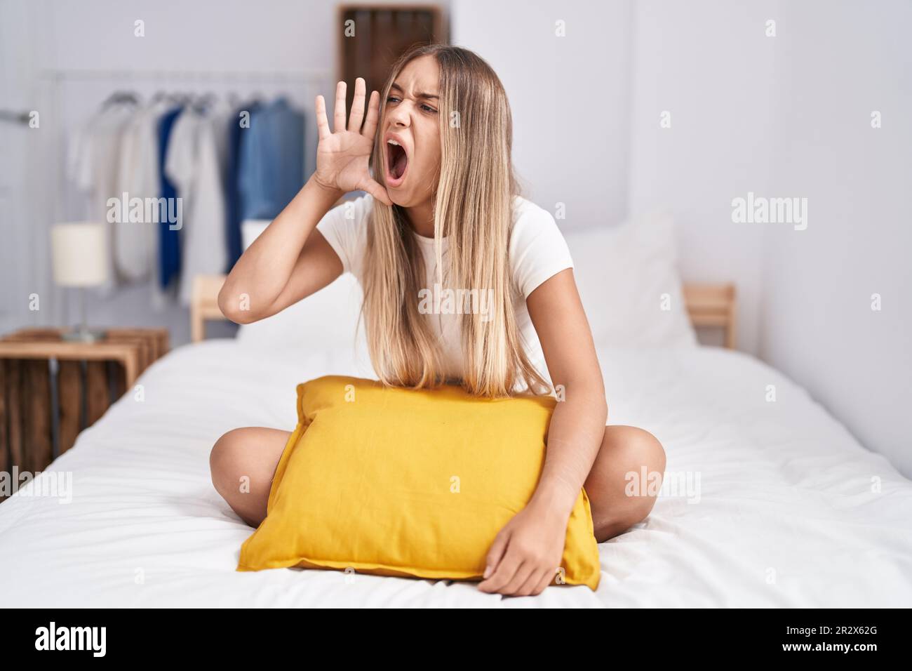 Young blonde woman sitting on the bed with pillow at home shouting and ...