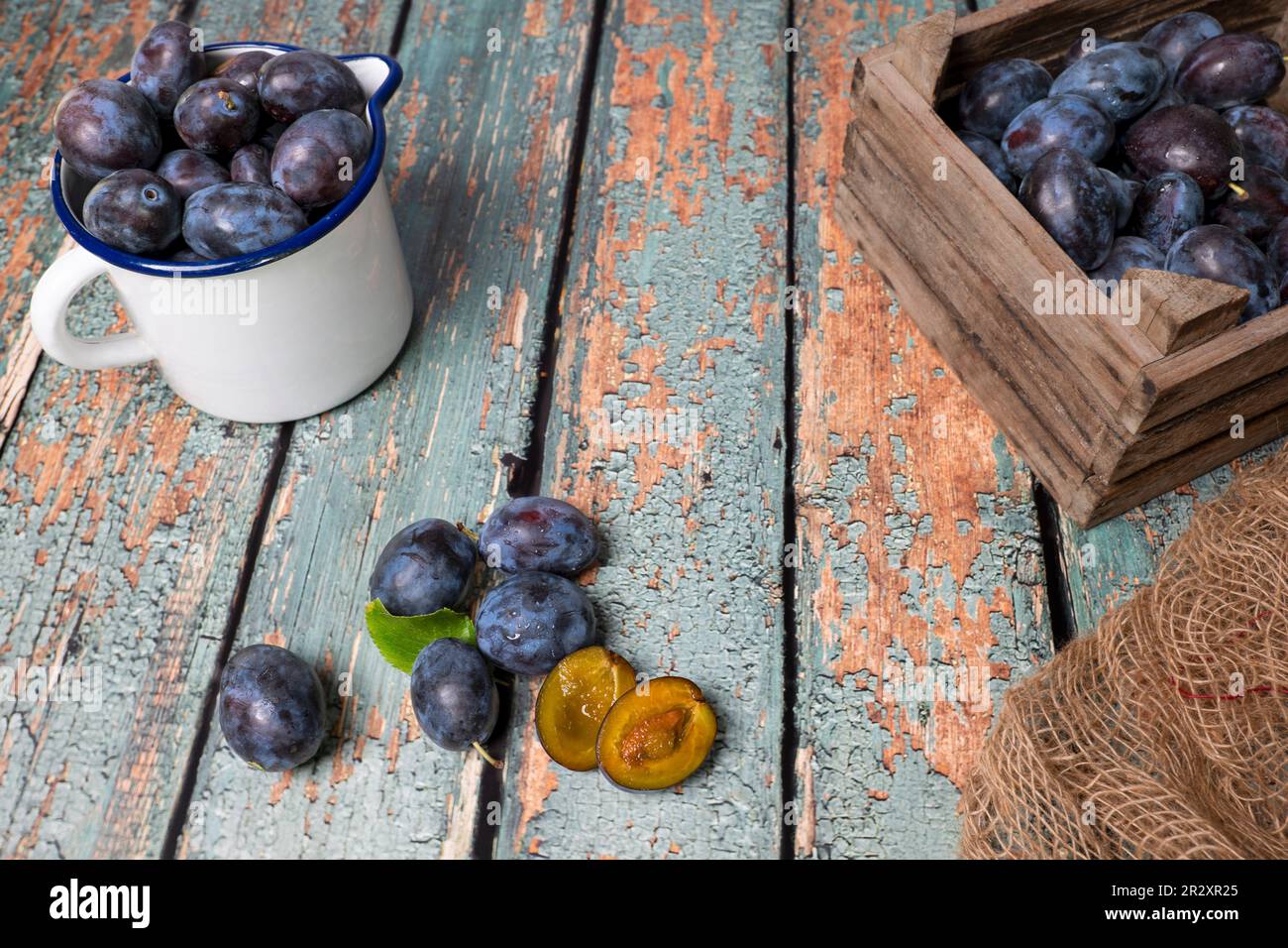 An enamel jug and a wooden box full of freshly harvested plums on old rustic wooden tabletop. A plum cut open. Stock Photo