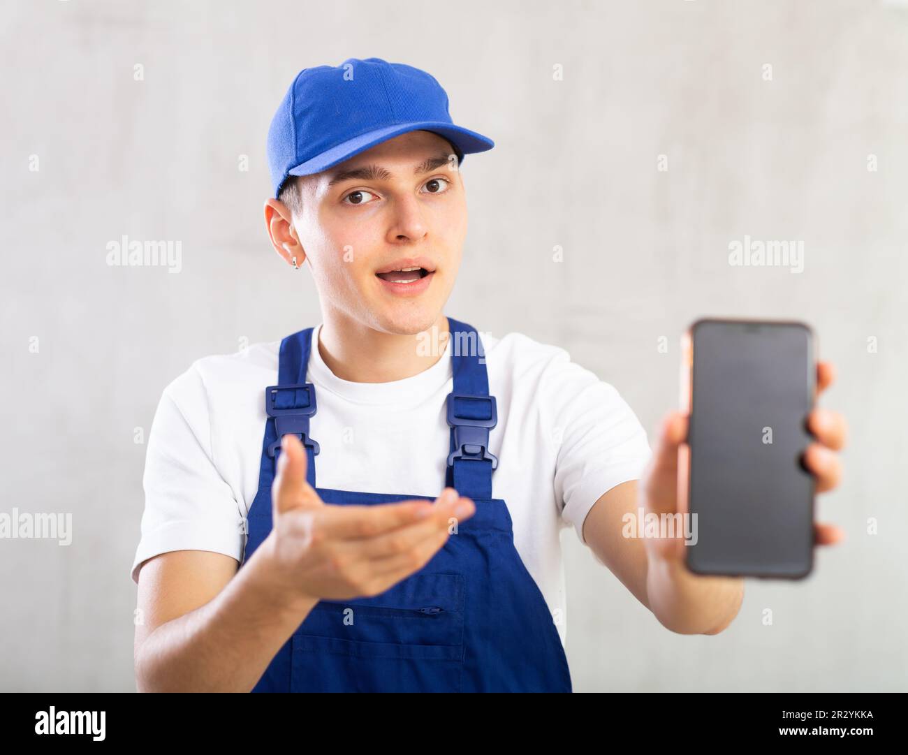 Man in jumpsuit and cap shows dark empty mobile phone screen and points ...