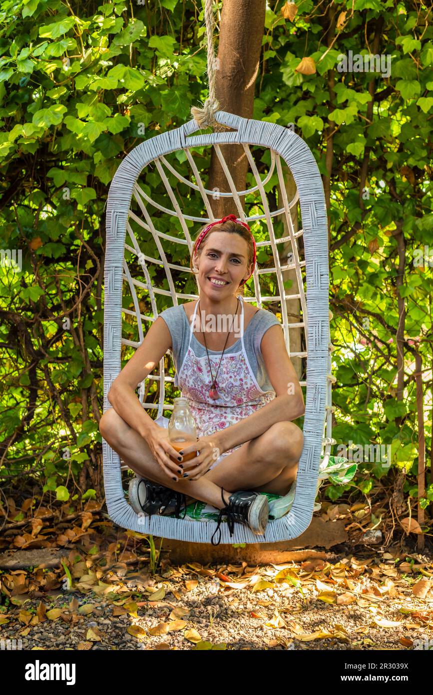 Smiling entrepreneur at her juice shop backyard resting on swing chair while drinking fresh detox juice Stock Photo