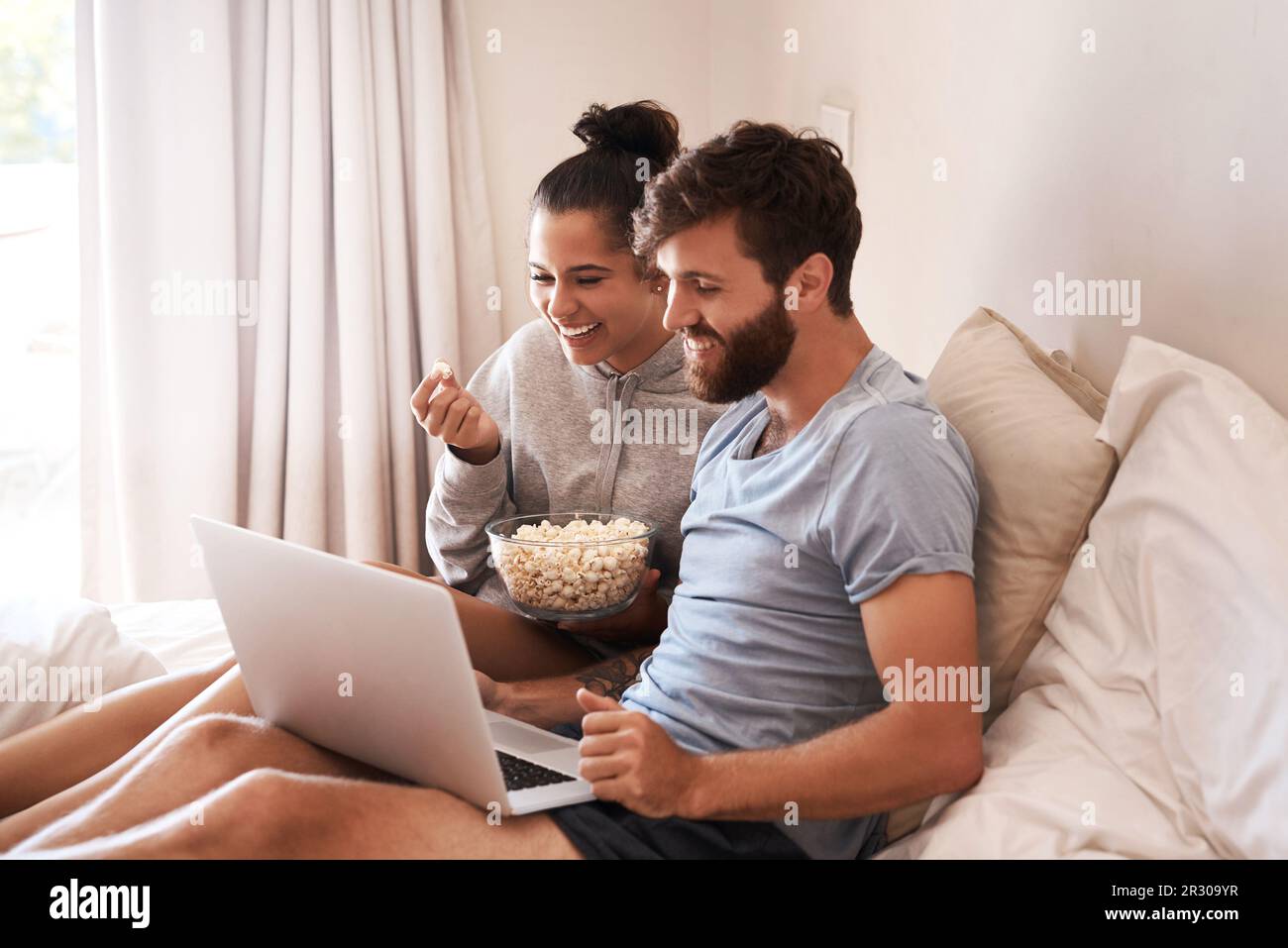 Watching movie, popcorn and couple in bed with laptop for streaming, food and internet. Subscription, happy and comic with man and woman at home for Stock Photo