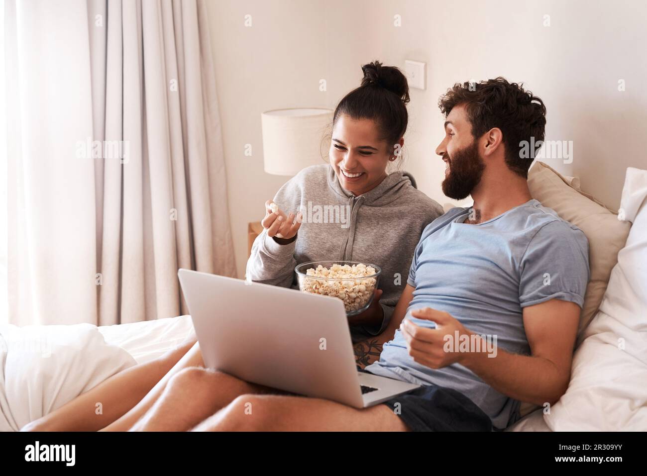 Laptop, watching movie and popcorn with laughing couple in bed for streaming, relax or internet. Subscription, happy or comic man and woman at home Stock Photo