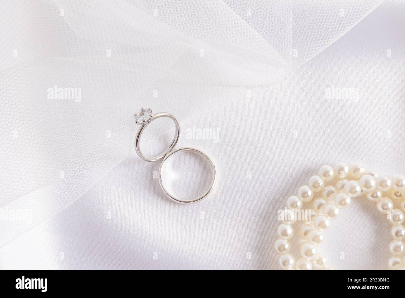 Two white gold rings with a diamond on the part of the bride's wedding veil and satin white textiles with pearl beads. Wedding composition Stock Photo