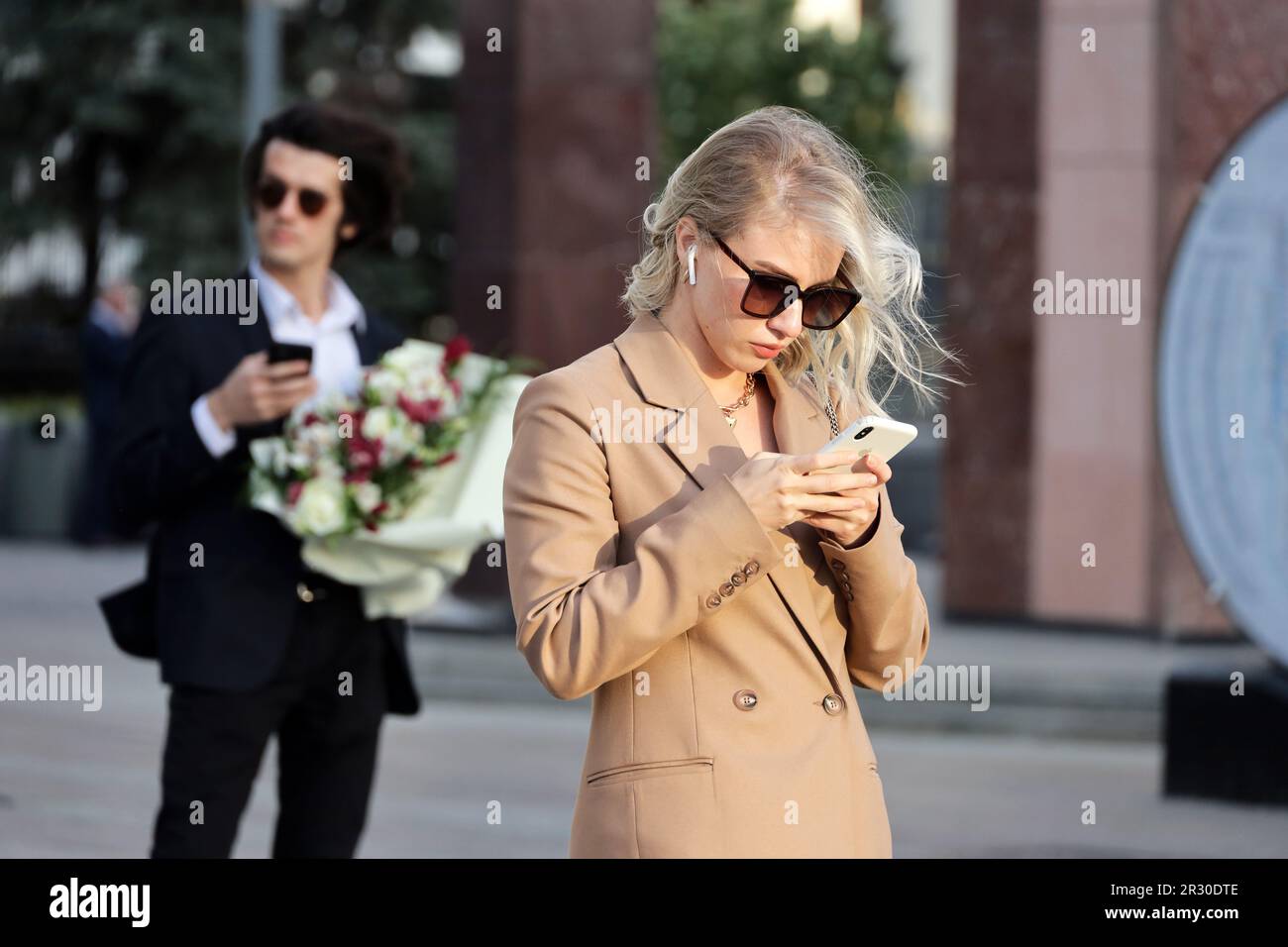 Blonde girl in sunglasses standing with smartphone in hands on city street on background of man with bouqet of flowers Stock Photo