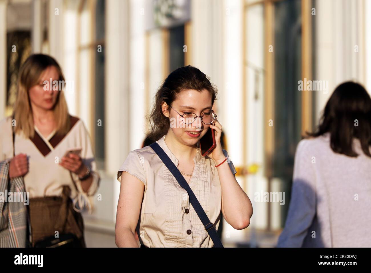 Happy girl in eyeglasses talking on smartphone while walking down street. Using mobile phone in spring city Stock Photo