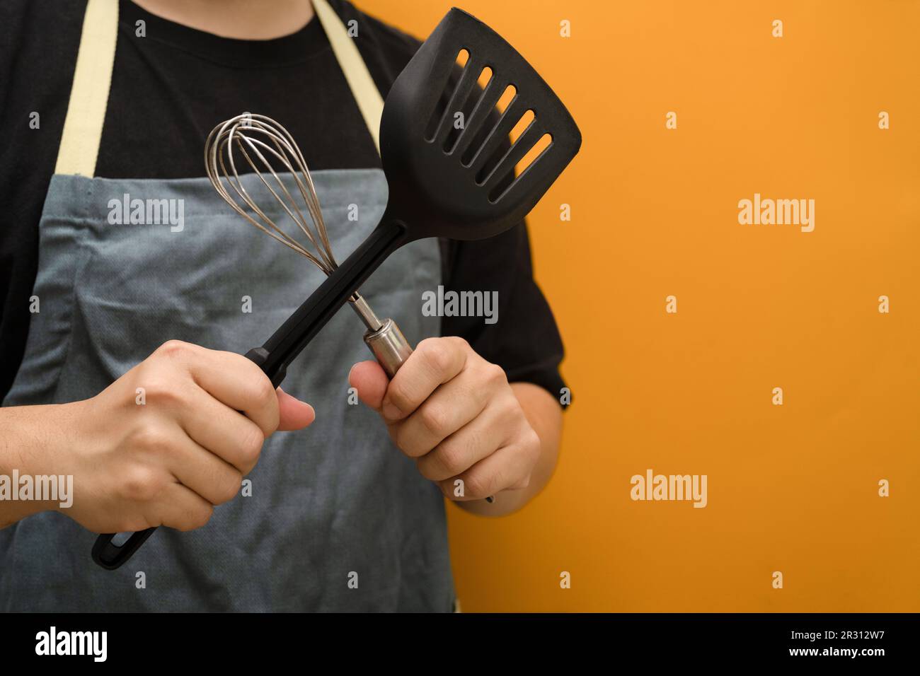 Man wearing apron holding whisks and kitchen spatula standing against yellow background with space Stock Photo
