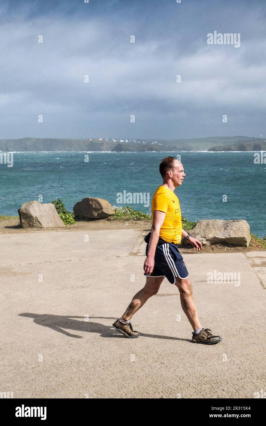 A man wearing shorts walking along the coast road on Towan Head overlooking Newquay Bay in Cornwall in the UK. Stock Photo