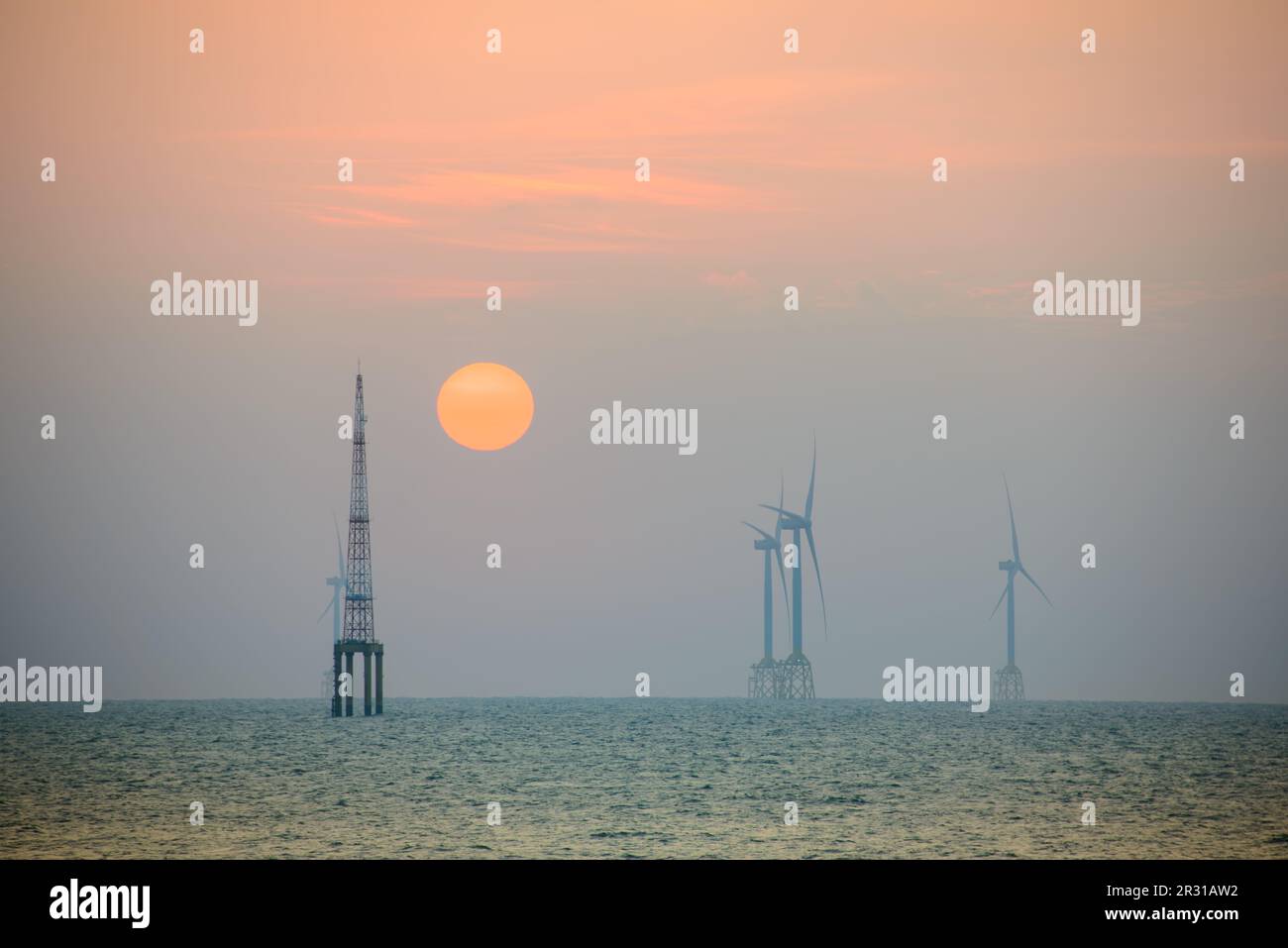 Fans of wind turbines spin over the sparkling sea. The orange-red sun at dusk. An offshore wind farm off the northwest coast of Taiwan. One of green p Stock Photo