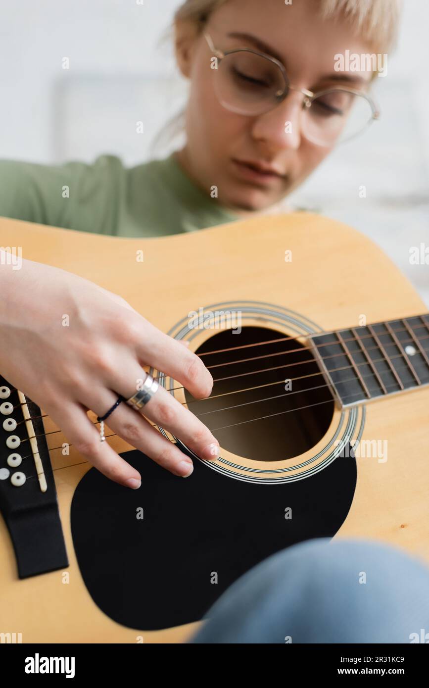 talanted young woman in glasses with bangs, rings on fingers playing acoustic guitar and sitting in modern living room, learning music, skill developm Stock Photo