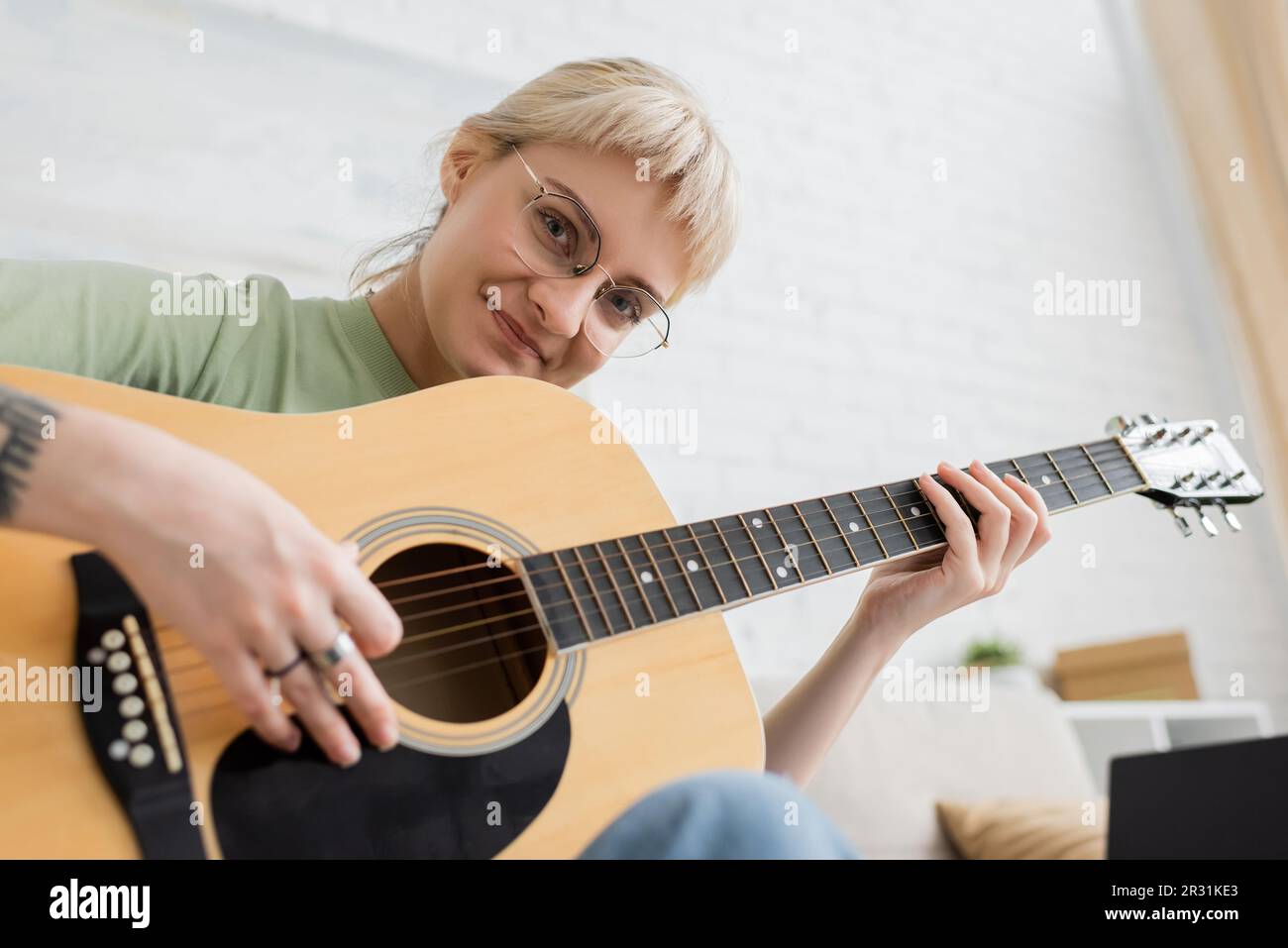 cheerful young woman in glasses with bangs and tattoo on hand playing acoustic guitar and looking at camera while sitting in modern living room, learn Stock Photo