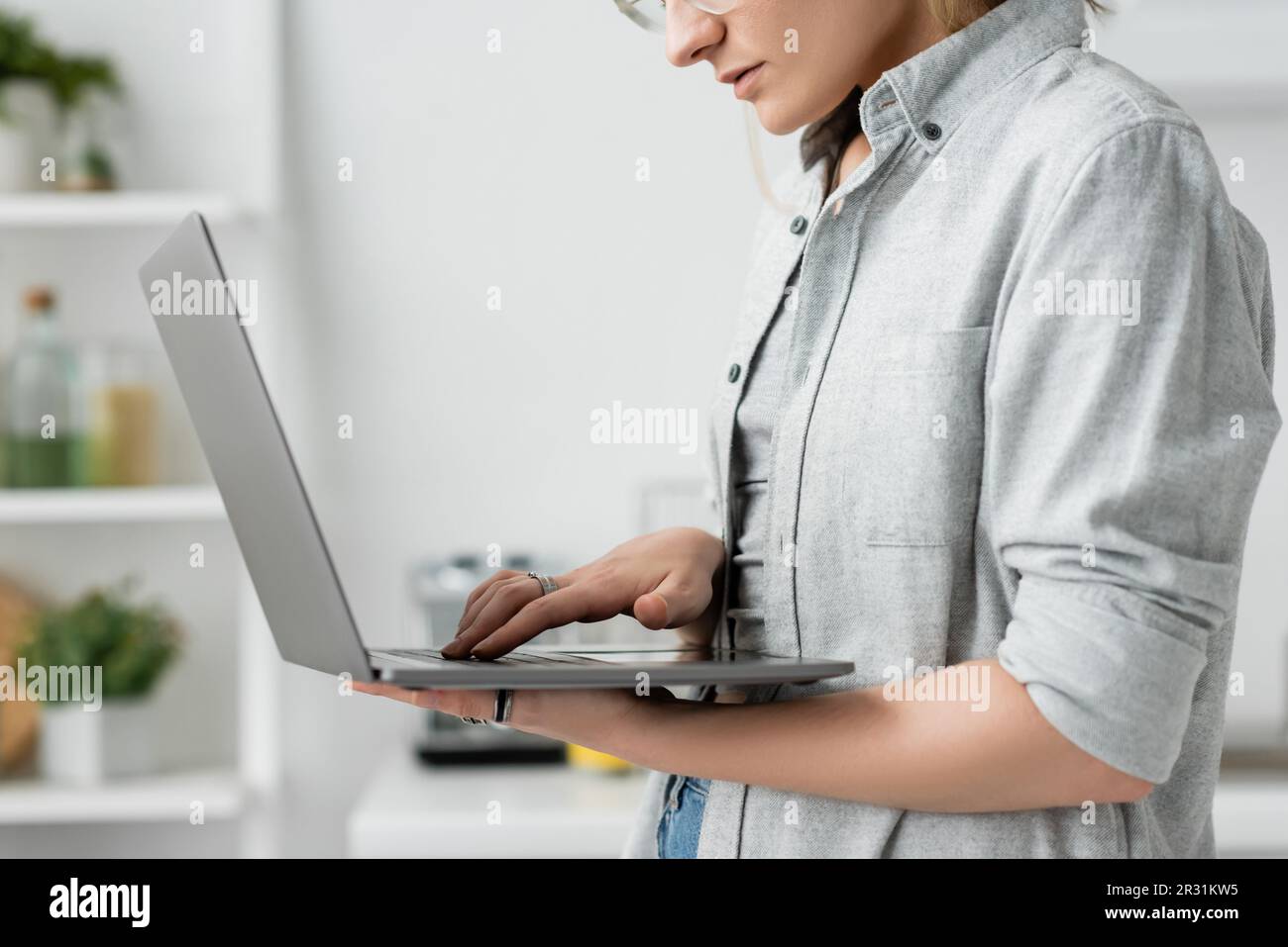 cropped view of focused young woman in grey shirt holding and using laptop in white and modern kitchen, blurred background, remote lifestyle, freelanc Stock Photo