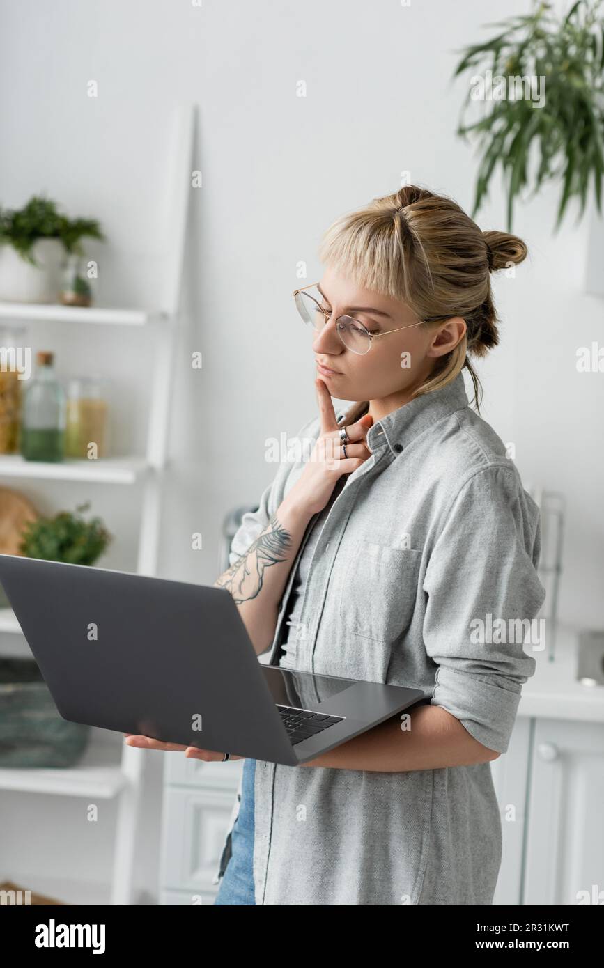 pensive young woman in eyeglasses, with tattoo and bangs using laptop while working from home in white and modern kitchen, blurred background, remote Stock Photo