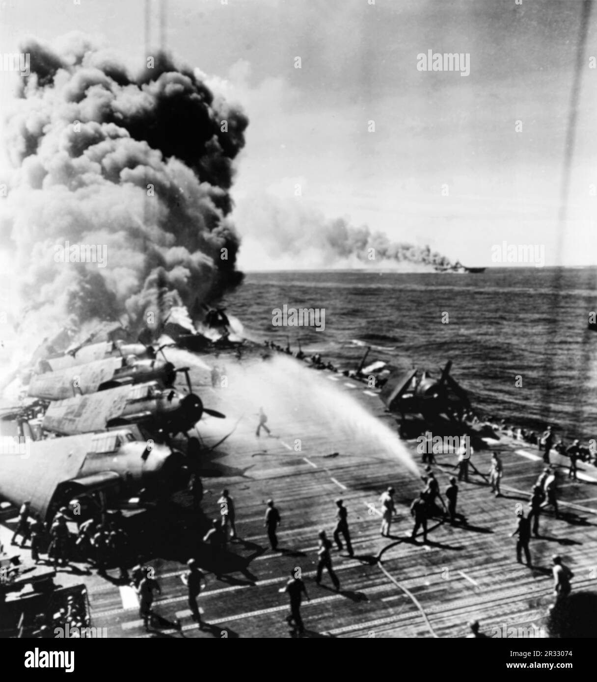 The U.S. Navy light aircraft carrier USS Belleau Wood (CVL-24) burning aft after she was hit by a Kamikaze, while operating off Luzon, Philippines, on 30 October 1944. Flight deck crewmen are moving undamaged Grumman TBM Avenger planes of Torpedo Squadron 21 (VT-21) away from the flames as others fight the fires. USS Franklin (CV-13), also hit during this Kamikaze attack, is afire in the distance. Stock Photo