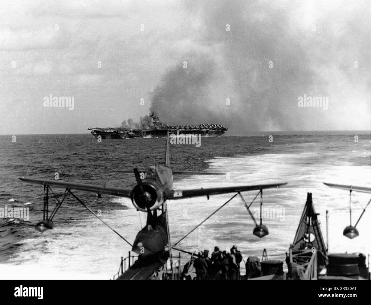 USS Ticonderoga (CV-14) afire after she was hit by a 'Kamikaze' attack off Formosa, 21 January 1945. Photographed from USS Miami (CL-89). A Vought OS2U 'Kingfisher' floatplane is on the cruiser's starboard catapult, in the foreground. When Japan was facing defeat in late 1944 it chose to destroy US ships with suicide bombings, known as Kamikaze.These attacks were a potent physical and psychological weapon and sunk a total of 47 ships at a cost of more than 3000 pilots and planes. By late 1944 the US Navy was large enough that the losses were insignificant and they did not alter the course of t Stock Photo