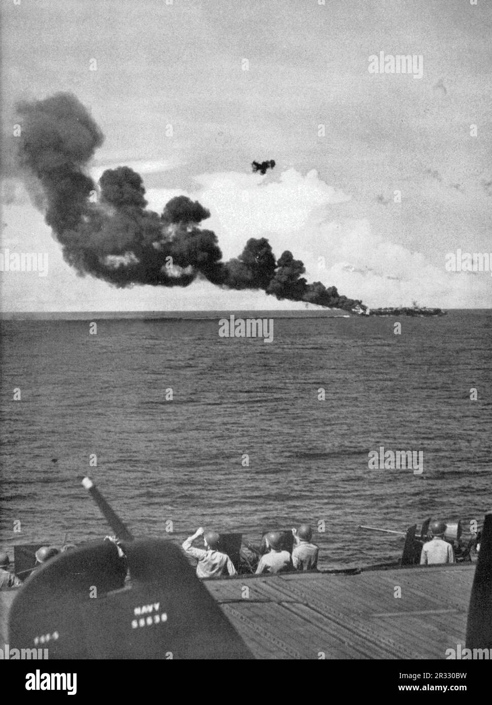 The U.S. Navy light aircraft carrier USS Belleau Wood (CVL-24) burning aft after she was hit by a Kamikaze, while operating off Luzon, Philippines, on 30 October 1944. Members of the Marine Detachment on board USS Enterprise (CV-6) shield their eyes from the sun as they scan the skies for kamikaze aircraft diving out of the sun. In the foreground is the tail of a Grumman F6F-5 Hellcat of Fighting Squadron 20 (VF-20). Stock Photo