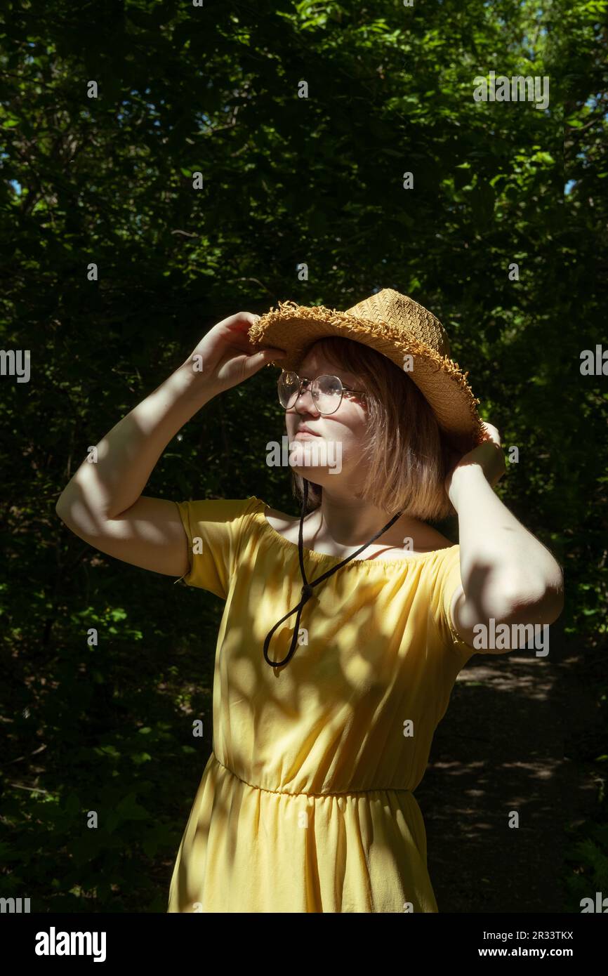 Beautiful Asian girl with glasses, straw hat and yellow dress in the forest in summer, beautiful shadows, sunlight, portrait. Dreamy young teen girl. Stock Photo