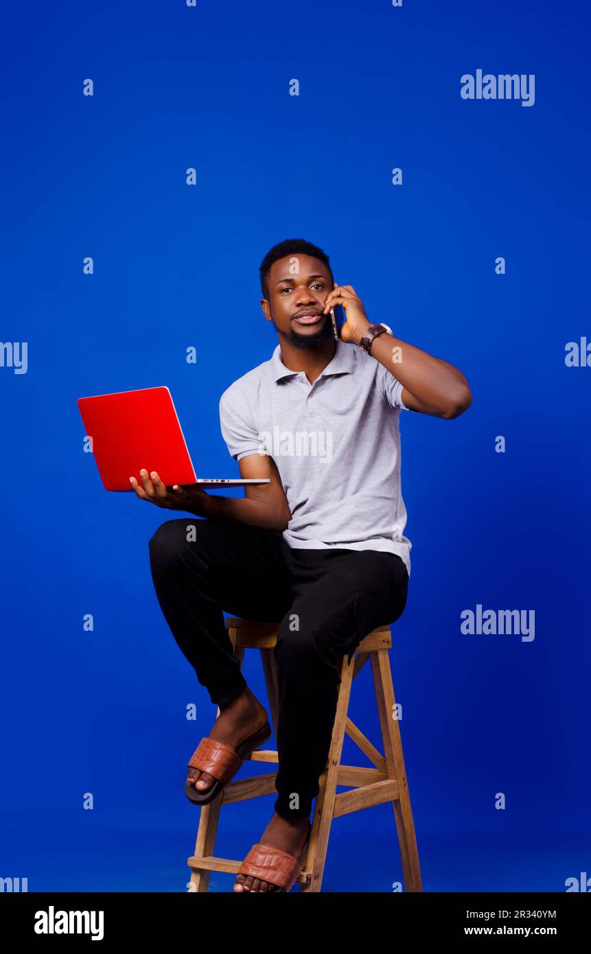 African man sitting on a chair while carrying laptop and using his mobile phone looking at the camera, isolated on a blue background Stock Photo