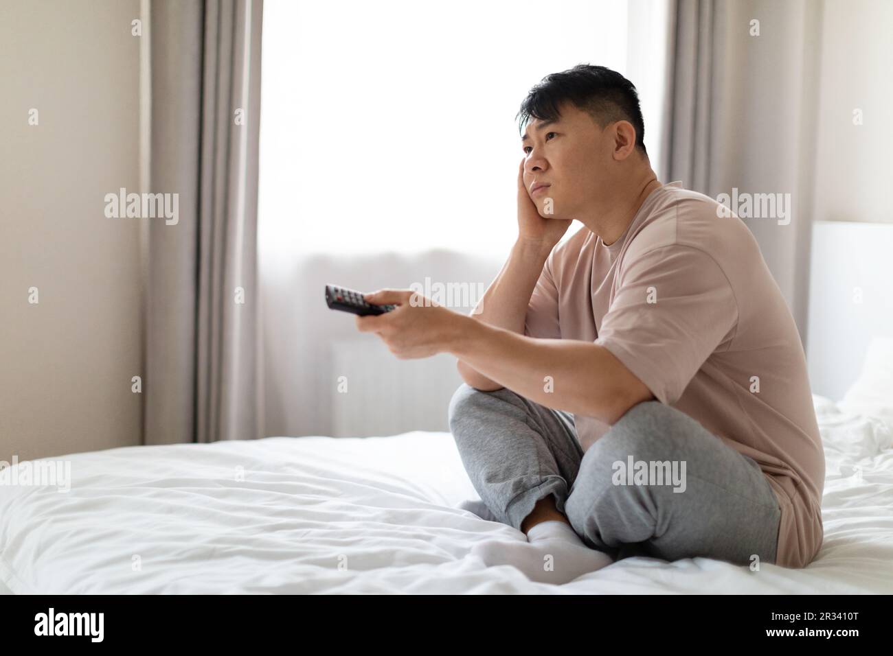 Unhappy middle aged asian man sitting on bed, watching TV Stock Photo
