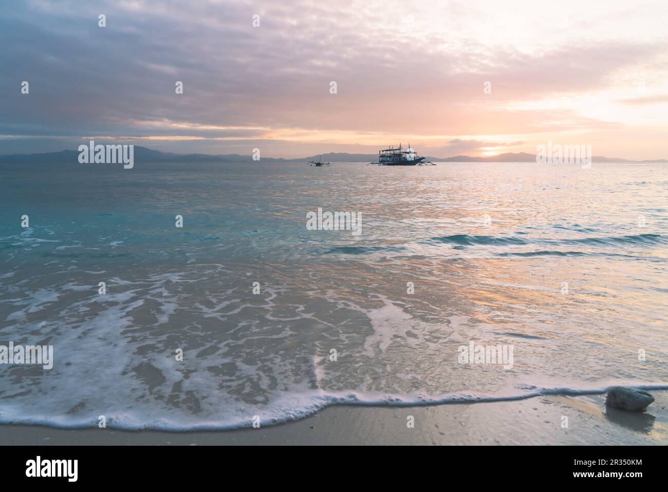 Scenic orange colored sunset on the beach with turquoise ocean surf and traditional boat on the horizon, Palawan, Philippines Stock Photo