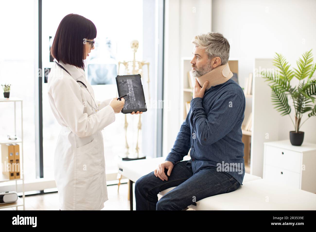 Caucasian lady holding tablet with CT scan image on screen while talking to male in neck brace sitting on exam couch. Young orthopedist analyzing thoracic vertebrae on digital report in clinic. Stock Photo