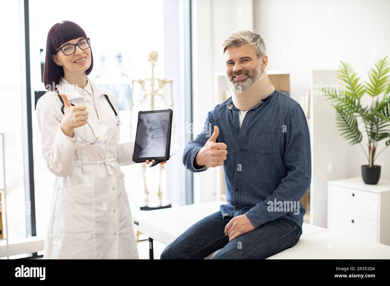 Cheerful young woman with digital tablet and smiling mature man in cervical collar posing in doctor's office. Therapist examining spine x-rays while happy patient sitting on exam couch indoors. Stock Photo