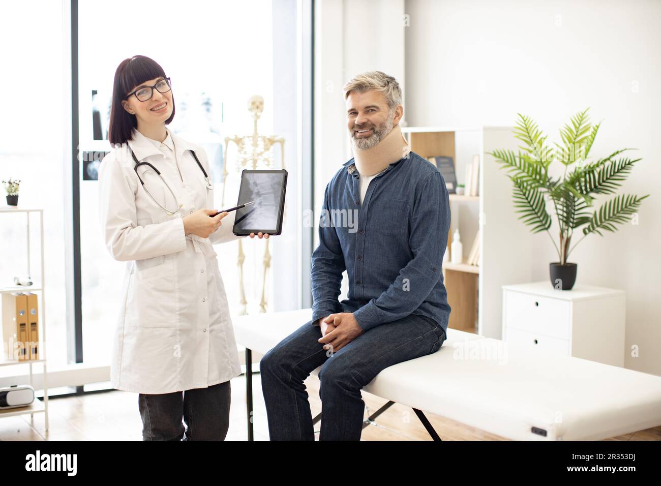 Cheerful young woman with digital tablet and smiling mature man in cervical collar posing in doctor's office. Therapist examining spine x-rays while happy patient sitting on exam couch indoors. Stock Photo