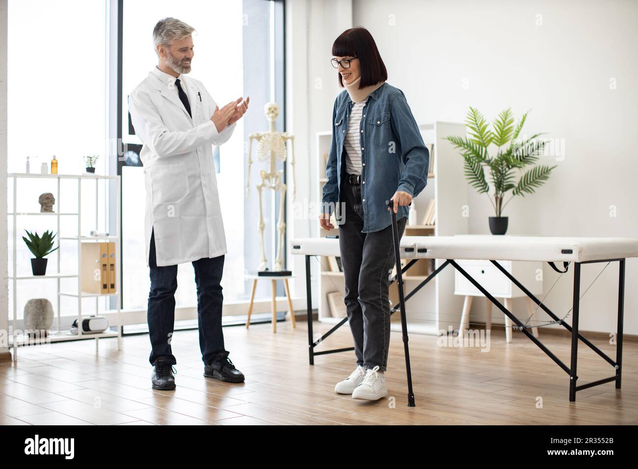 Cheerful doctor in white coat clapping hands while lady in C collar standing without