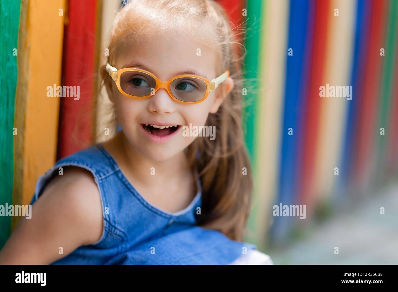 little girl with down syndrome wearing glasses for vision sits near a colored fence in a children's town in summer. High quality photo Stock Photo
