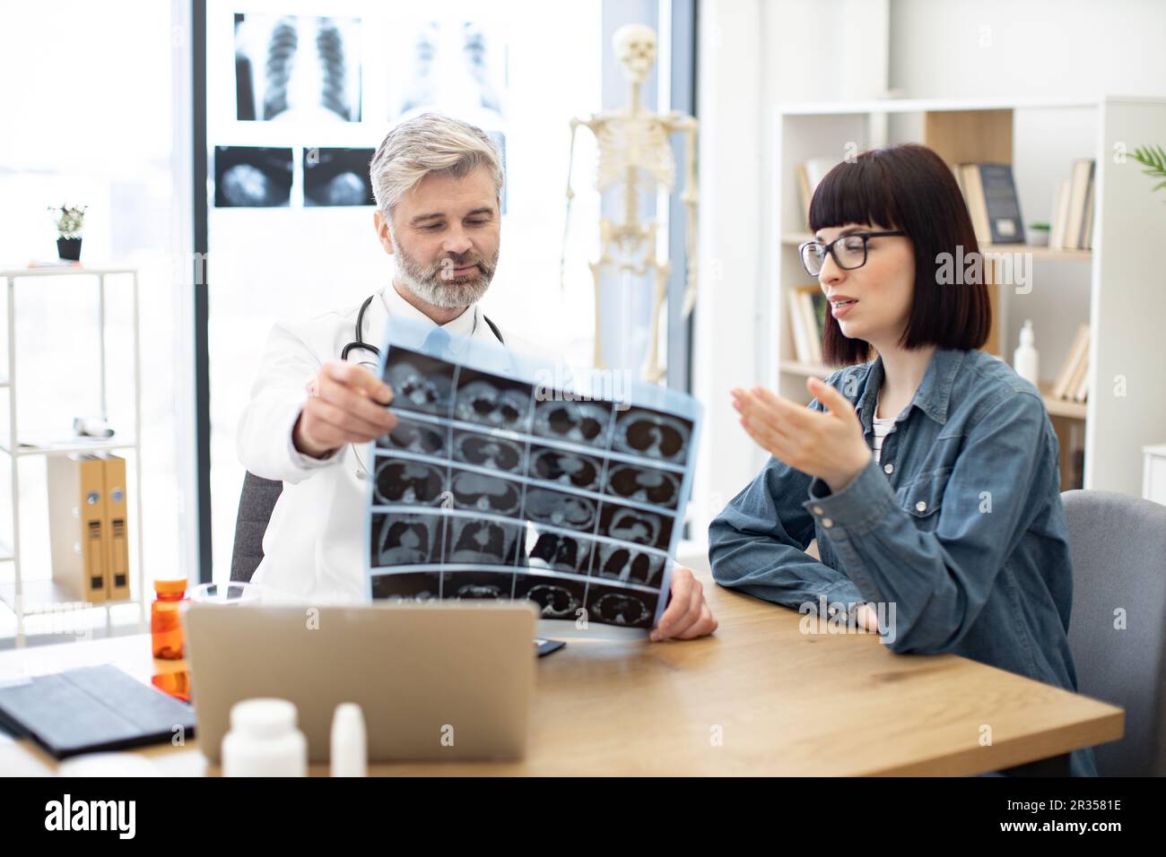 Mindful gray-haired male in lab coat holding MRI scans while brunette lady sitting next to him at desk in hospital. Family doctor examining diagnostic test results while developing treatment plan. Stock Photo