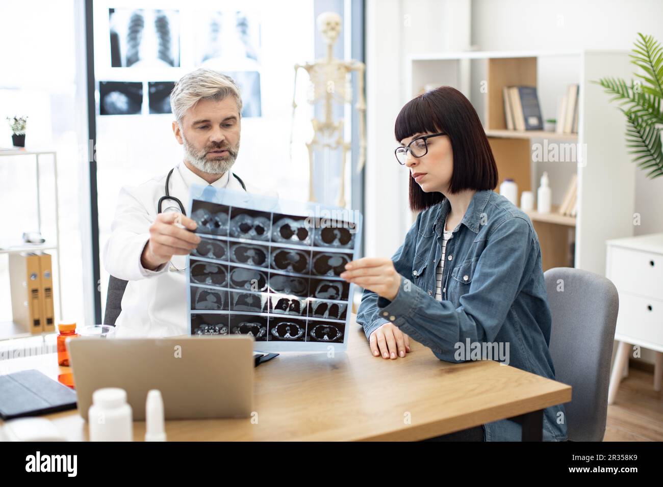 Mindful gray-haired male in lab coat holding MRI scans while brunette lady sitting next to him at desk in hospital. Family doctor examining diagnostic test results while developing treatment plan. Stock Photo
